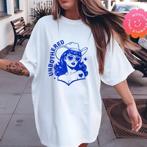 May include: White oversized t-shirt with a blue graphic of a woman wearing a cowboy hat and sunglasses. The graphic says "UNBOTHERED" in blue letters.