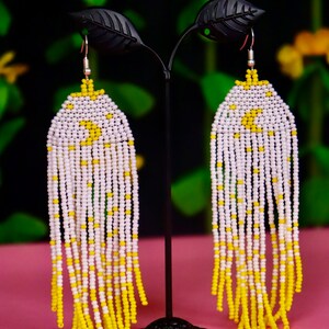 Puede incluir: Un par de aretes largos de cuentas con un esquema de color blanco y amarillo. Los aretes están colgados de un soporte negro.