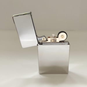 May include: A polished, silver-toned lighter with the lid open, displaying the internal mechanism. The lighter has a rectangular shape and a hinged lid. The wick and flint components are visible. The lighter is presented against a simple white backdrop.