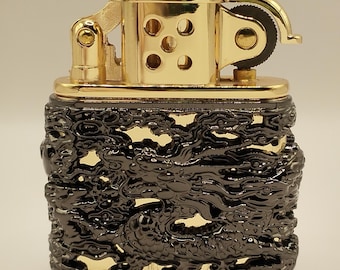 May include: A rectangular metal lighter featuring a polished gold-tone top mechanism with a flint wheel and a black, openwork outer casing decorated with a detailed, raised dragon relief pattern that reveals the gold-tone surface underneath.