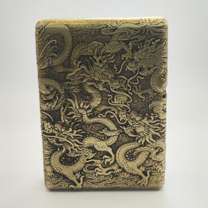 May include: A rectangular, antique brass-colored lighter with an intricate dragon design carved into its surface. The dragons are depicted in a detailed, embossed style, creating a textured appearance. The lighter's design is a classic, collectible item.