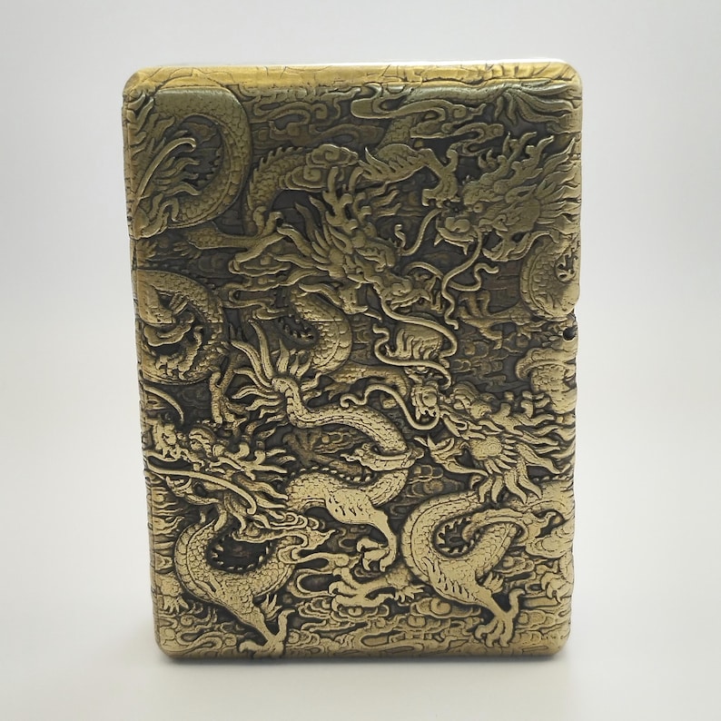 May include: A rectangular, antique brass-colored lighter with an intricate dragon design carved into its surface. The dragons are depicted in a detailed, embossed style, creating a textured appearance. The lighter's design is a classic, collectible item.