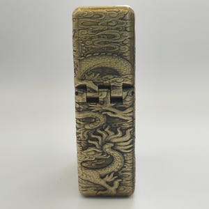 May include: A gold-colored lighter with intricate dragon and cloud relief carvings. The lighter's design features a detailed dragon motif, with a hinged top and a rectangular shape. The overall aesthetic is vintage and ornate.
