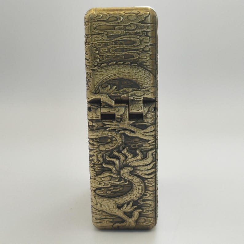 May include: A gold-colored lighter with intricate dragon and cloud relief carvings. The lighter's design features a detailed dragon motif, with a hinged top and a rectangular shape. The overall aesthetic is vintage and ornate.