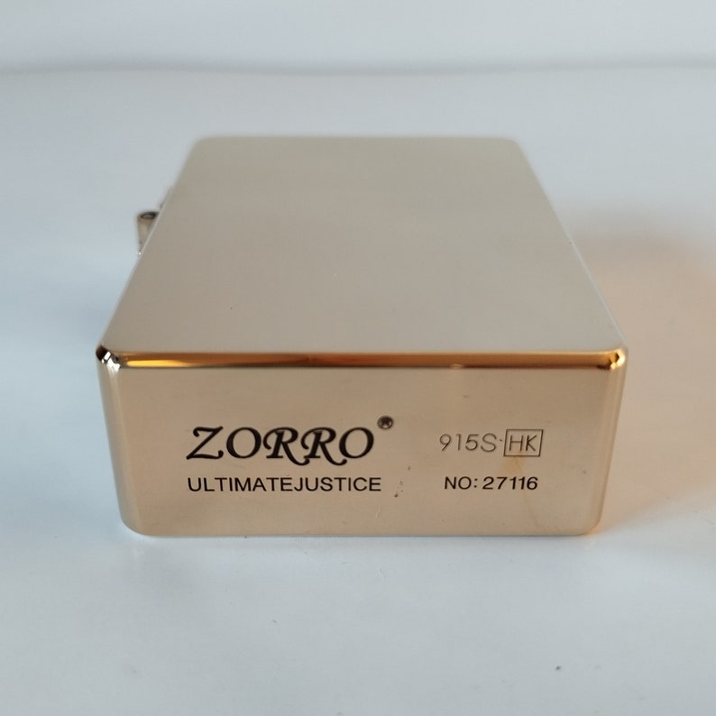 Op de afbeelding: Gouden aansteker met de tekst "ZORRO ULTIMATEJUSTICE 915S-HK NO:27116" gedrukt op de onderkant.