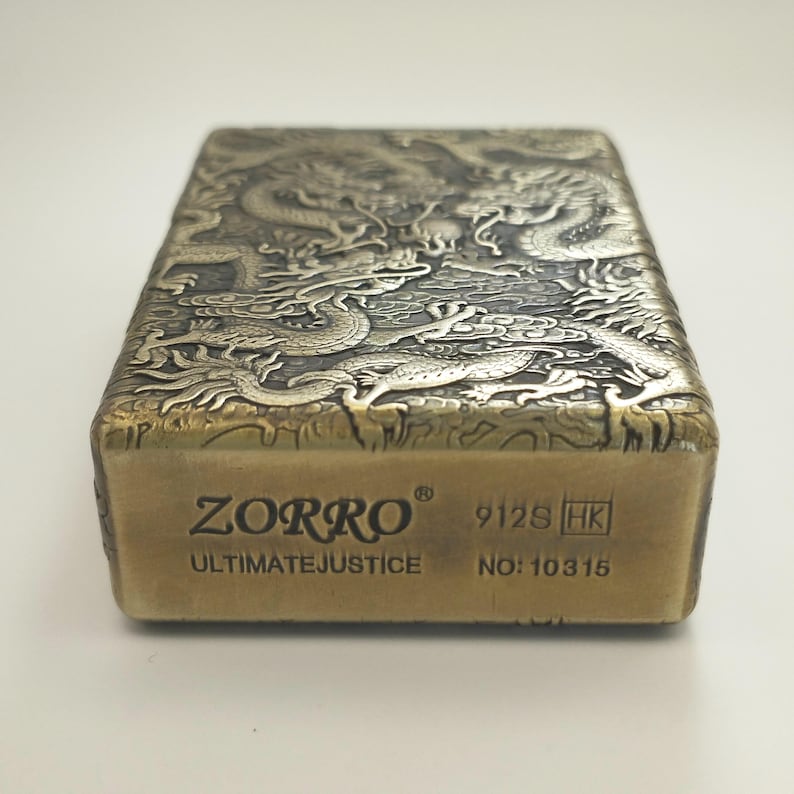 May include: A brass-colored ZORRO lighter with an intricate dragon design engraved on the top. The lighter has the text "ZORRO ULTIMATE JUSTICE" and the number "10315" printed on the front.
