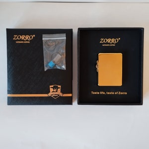 Op de afbeelding: Een gouden aansteker in een zwarte doos met de woorden "Zorro Ultimate Justice" en "Taste life, taste of Zorro".