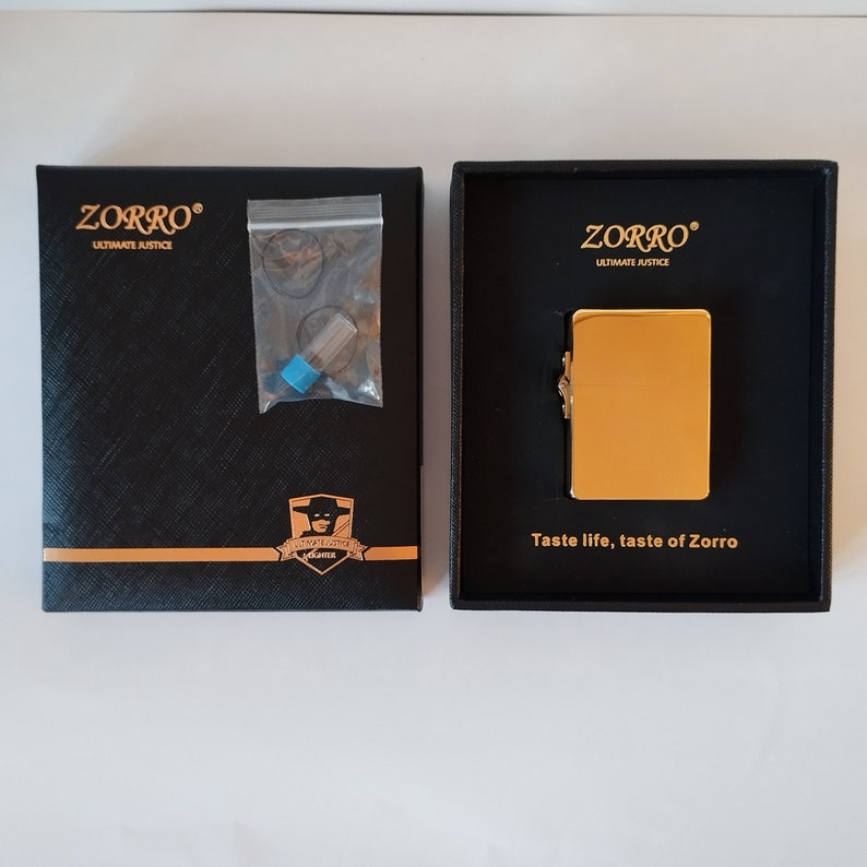 Op de afbeelding: Een gouden aansteker in een zwarte doos met de woorden "Zorro Ultimate Justice" en "Taste life, taste of Zorro".