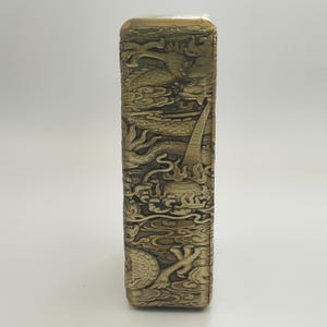May include: A rectangular, antique-style lighter with intricate dragon and cloud relief carvings. The lighter has a metallic gold finish and detailed artwork across its surface, showcasing a classic design.
