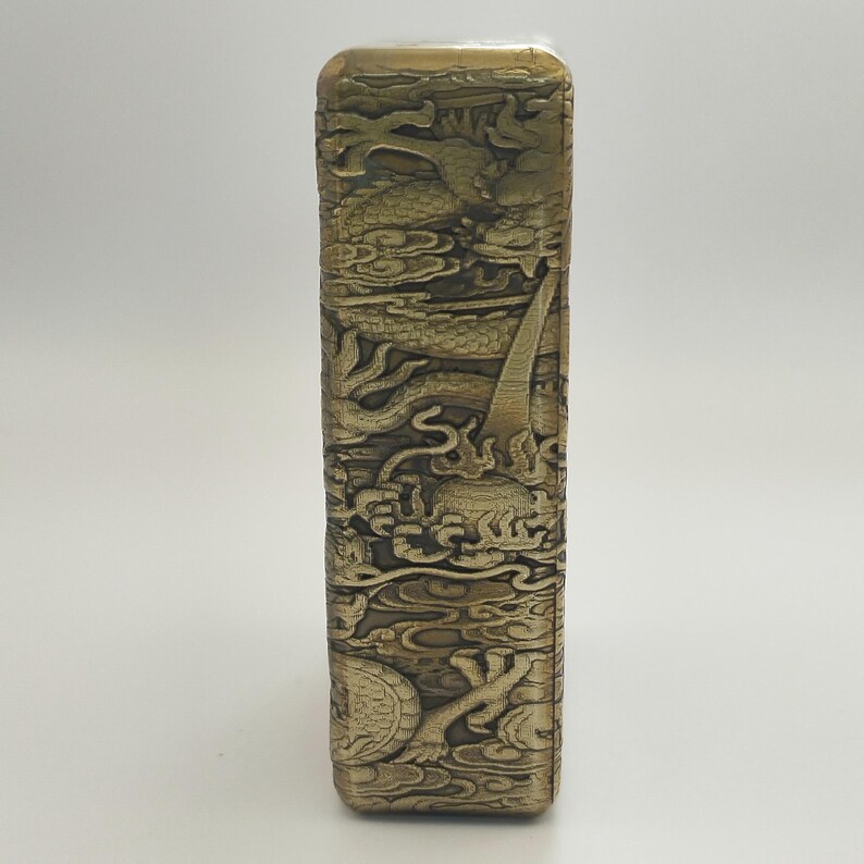 May include: A rectangular, antique-style lighter with intricate dragon and cloud relief carvings. The lighter has a metallic gold finish and detailed artwork across its surface, showcasing a classic design.