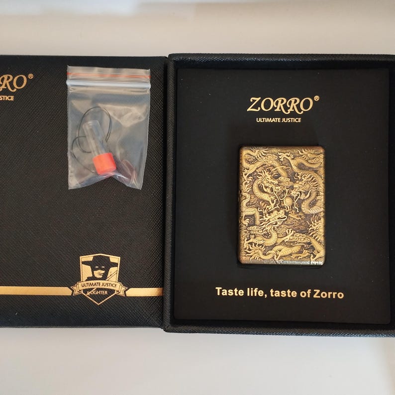 May include: A ZORRO lighter with a dragon design, presented in a black box. The lighter has a detailed, embossed dragon pattern in a bronze color. The box includes the text "ZORRO ULTIMATE JUSTICE" and "Taste life, taste of Zorro."