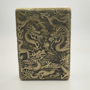 May include: A rectangular, antique-style lighter with a detailed relief of dragons. The lighter has a gold finish and the dragons are intricately carved, creating a textured surface. The design features multiple dragons intertwined.