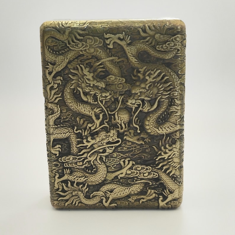 May include: A rectangular, antique-style lighter with a detailed relief of dragons. The lighter has a gold finish and the dragons are intricately carved, creating a textured surface. The design features multiple dragons intertwined.