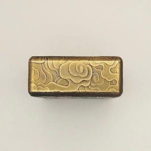 May include: A rectangular, brass-colored box with a cloud design. The box has rounded edges and a detailed, embossed pattern of swirling clouds across the top surface. The metal has a slightly aged appearance.