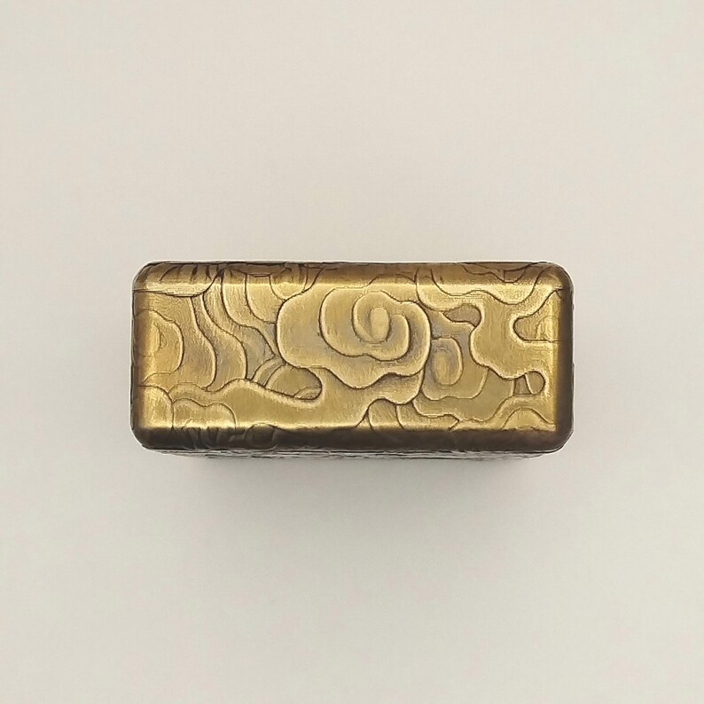 May include: A rectangular, brass-colored box with a cloud design. The box has rounded edges and a detailed, embossed pattern of swirling clouds across the top surface. The metal has a slightly aged appearance.