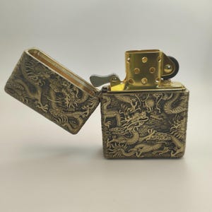 May include: A brass-colored lighter with an open lid, revealing a gold-toned interior. The lighter's exterior is intricately embossed with a dragon design. The lighter is a square shape and has a gold-colored top.