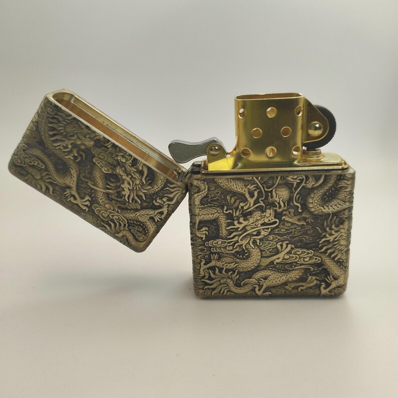 May include: A brass-colored lighter with an open lid, revealing a gold-toned interior. The lighter's exterior is intricately embossed with a dragon design. The lighter is a square shape and has a gold-colored top.