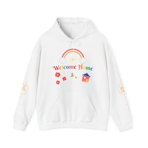 Può includere: Felpa con cappuccio bianca con un arcobaleno, sole, nuvole, fiori, api e un disegno di una casa. Il testo "Welcome Home" è stampato sul davanti della felpa.