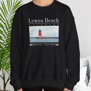 May include: Black crew neck sweatshirt with a graphic of a red and white lighthouse on an island, with the text "Lewes Beach Delaware" and "Est. 1631" in white. The sweatshirt is a casual style.