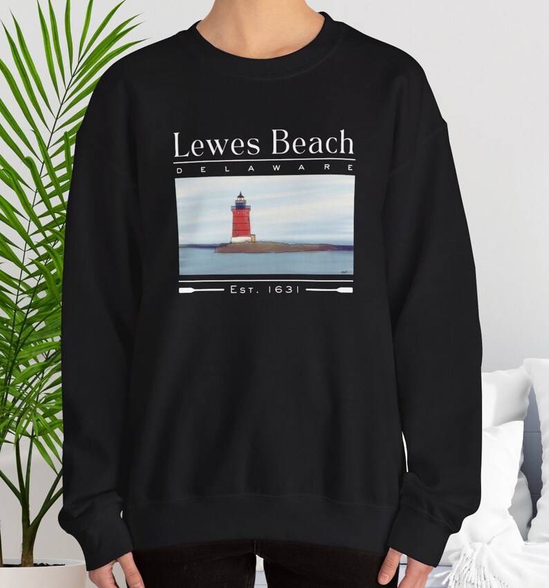 May include: Black crew neck sweatshirt with a graphic of a red and white lighthouse on an island, with the text "Lewes Beach Delaware" and "Est. 1631" in white. The sweatshirt is a casual style.
