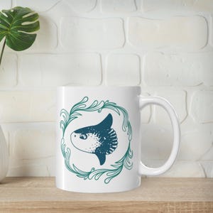 May include: White ceramic mug with a blue and white illustration of a mola mola fish surrounded by green leafy vines.