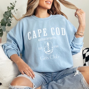 May include: A light blue sweatshirt with the words "CAPE COD MASSACHUSETTS" in white block letters. Below the text is a white graphic of a lighthouse and the words "EST. 1708 Girls Club". The sweatshirt is a crew neck style.