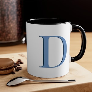 May include: A white ceramic mug with a black rim and handle. The mug has a blue letter "D" printed on the front.