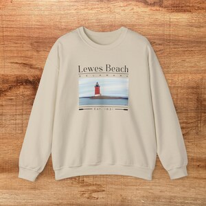 May include: Cream-coloured sweatshirt with a graphic of a red lighthouse on a small island. The text reads "Lewes Beach Delaware Est. 1631". The sweatshirt has long sleeves and a crew neck.