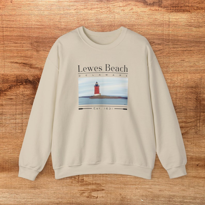 May include: Cream-coloured sweatshirt with a graphic of a red lighthouse on a small island. The text reads "Lewes Beach Delaware Est. 1631". The sweatshirt has long sleeves and a crew neck.