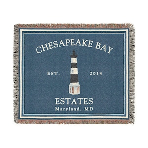 May include: A blue woven throw blanket with a white border and fringed edges. The center features a lighthouse graphic and text that reads "CHESAPEAKE BAY ESTATES Maryland, MD" and "EST. 2014".