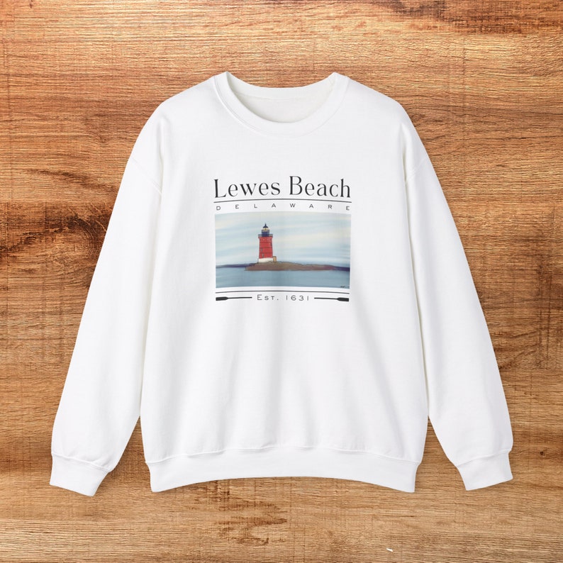 May include: White sweatshirt with a graphic of a red lighthouse on an island, the text reads "Lewes Beach Delaware" and "Est. 1631". The sweatshirt has long sleeves and a crew neck.
