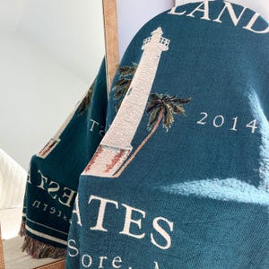 May include: A teal blue woven throw blanket with a white lighthouse, palm trees, and the text "Gates Island 2014" in white.