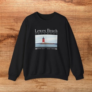 May include: Black sweatshirt with a graphic of a red lighthouse on a small island, with the words "Lewes Beach Delaware" and "Est. 1631" printed in white. The sweatshirt is made of a soft material.