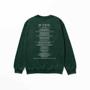 Pode incluir: Uma camisola verde escura com a palavra "RUNWAY" impressa nas costas, seguida de uma lista de cargos e nomes.
