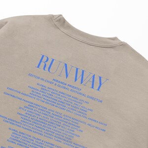 Pode incluir: Uma camiseta bege com a palavra "RUNWAY" impressa em azul. O texto "EDITOR-IN-CHIEF & GLOBAL EDITORIAL DIRECTOR" est&aacute; impresso abaixo da palavra "RUNWAY" em azul. O texto "MIRANDA PRIESTLY" est&aacute; impresso abaixo do t&iacute;tulo em azul. O texto abaixo do nome lista os nomes dos diretores de moda e seus t&iacute;tulos.