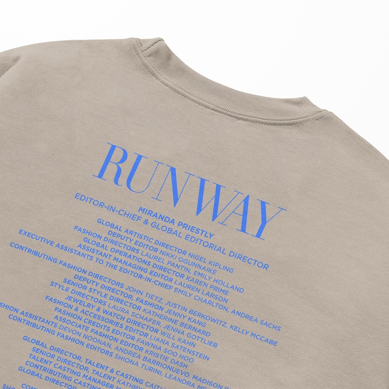 Pode incluir: Uma camiseta bege com a palavra "RUNWAY" impressa em azul. O texto "EDITOR-IN-CHIEF & GLOBAL EDITORIAL DIRECTOR" est&aacute; impresso abaixo da palavra "RUNWAY" em azul. O texto "MIRANDA PRIESTLY" est&aacute; impresso abaixo do t&iacute;tulo em azul. O texto abaixo do nome lista os nomes dos diretores de moda e seus t&iacute;tulos.