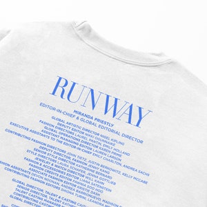 Pode incluir: Uma camiseta branca com o texto "RUNWAY" em letras azuis. O texto "EDITOR-IN-CHIEF & GLOBAL EDITORIAL DIRECTOR" &eacute; impresso abaixo do logotipo, seguido de uma lista de nomes e t&iacute;tulos, incluindo Miranda Priestly, Nigel Kipling e John Tietz.