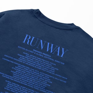 Pode incluir: Uma camiseta azul marinho com a palavra "RUNWAY" impressa em branco. A parte de tr&aacute;s da camiseta lista os nomes e os t&iacute;tulos da equipe editorial da revista.