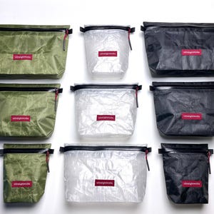 Op de afbeelding: Een collectie rechthoekige ritszakjes in olijfgroen, doorschijnend wit en zwart. Elk zakje is voorzien van een zwarte rits met een rood treklipje en een klein rood rechthoekig label met de witte tekst ultralightworks in het midden aan de voorzijde.
