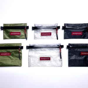 Könnte beinhalten: Eine Auswahl an Ultralightworks-Reißverschlusstaschen in verschiedenen Farben, darunter Blau, Grün, Transparent und Schwarz. Jede Tasche hat einen schwarzen Reißverschluss, einen roten Zuglasche und ein rotes rechteckiges Etikett mit dem Markennamen.