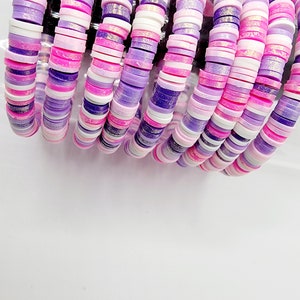 Peut inclure: Une pile de bracelets colorés faits de perles rondes, plates et en céramique. Les perles sont dans des tons de rose, de violet et de blanc avec un léger éclat.