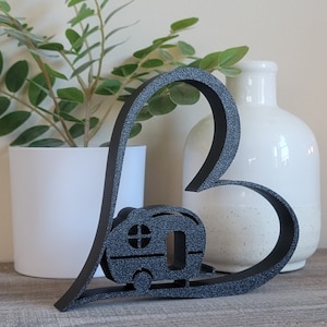 May include: Black wooden heart-shaped cutout with a silhouette of a camper van inside. The heart is sitting on a wooden surface in front of a potted plant and a white ceramic vase.