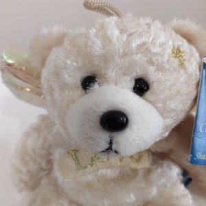 May include: A white plush teddy bear with a gold star on its chest and a gold ribbon around its neck that says "Love". The bear has iridescent wings and is a small, cuddly toy.
