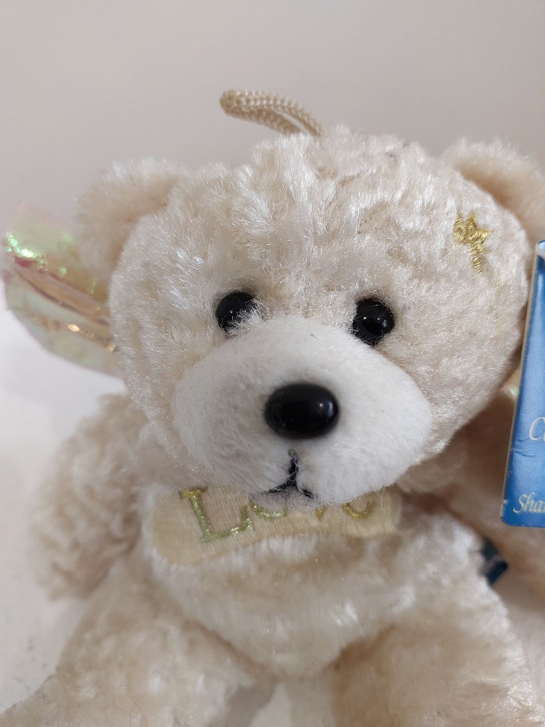 May include: A white plush teddy bear with a gold star on its chest and a gold ribbon around its neck that says "Love". The bear has iridescent wings and is a small, cuddly toy.