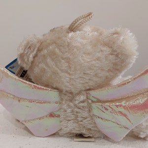 May include: A beige plush teddy bear with iridescent wings. The bear has a tag that says "To:" and a small tag with the word "TY".