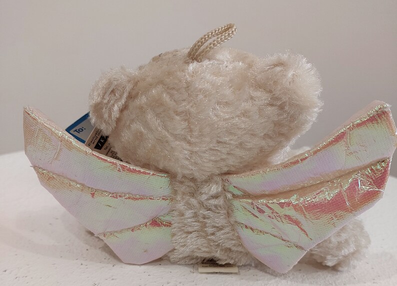 May include: A beige plush teddy bear with iridescent wings. The bear has a tag that says "To:" and a small tag with the word "TY".