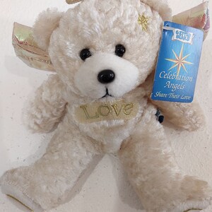May include: A white plush teddy bear with a gold halo and wings. The bear has a gold star on its chest and a gold "Love" inscription. The bear is holding a blue and white tag that reads "Gifted Bears Celebration Angels Share Their Love".