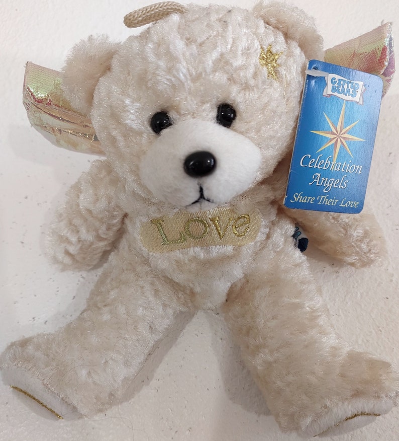 May include: A white plush teddy bear with a gold halo and wings. The bear has a gold star on its chest and a gold "Love" inscription. The bear is holding a blue and white tag that reads "Gifted Bears Celebration Angels Share Their Love".