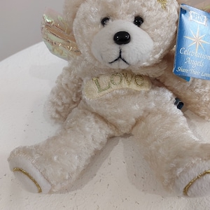 May include: A white plush teddy bear with a gold halo and wings. The bear has a gold "Love" inscription on its chest and a blue tag that reads "Celebration Angels Share Their Love".