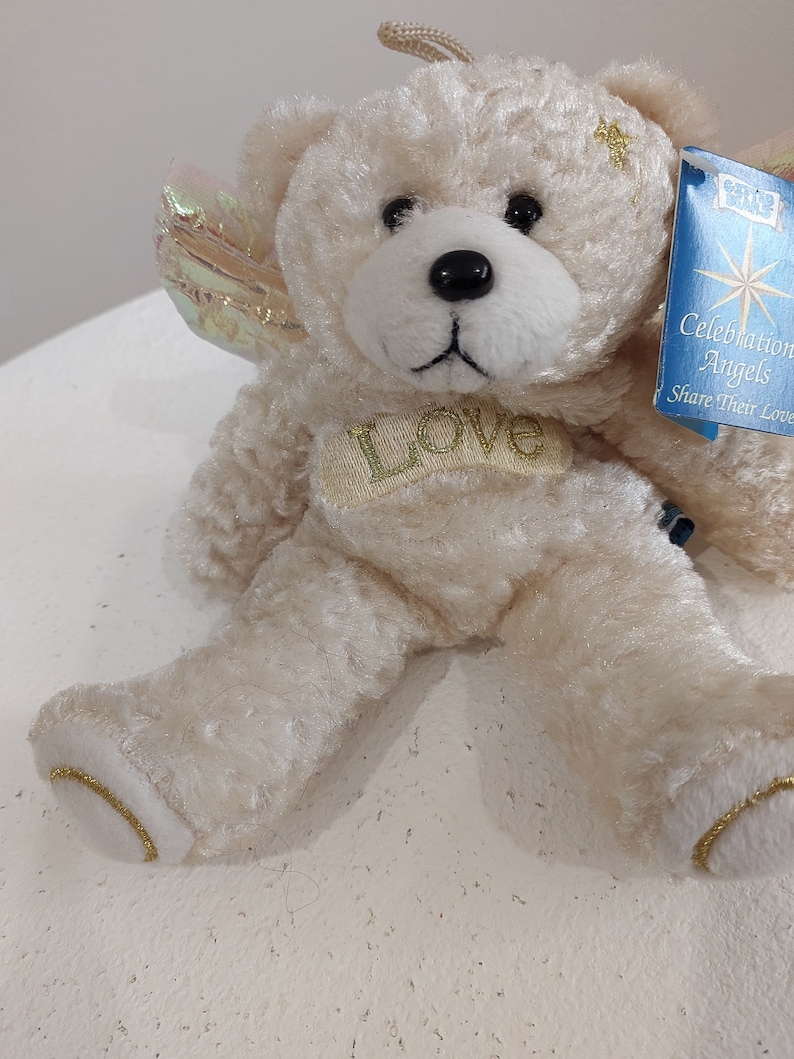 May include: A white plush teddy bear with a gold halo and wings. The bear has a gold "Love" inscription on its chest and a blue tag that reads "Celebration Angels Share Their Love".